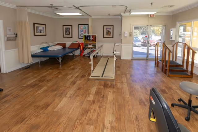 Spacious rehabilitation room with parallel bars, a therapy bed, exercise equipment and a small practice staircase near the entrance.