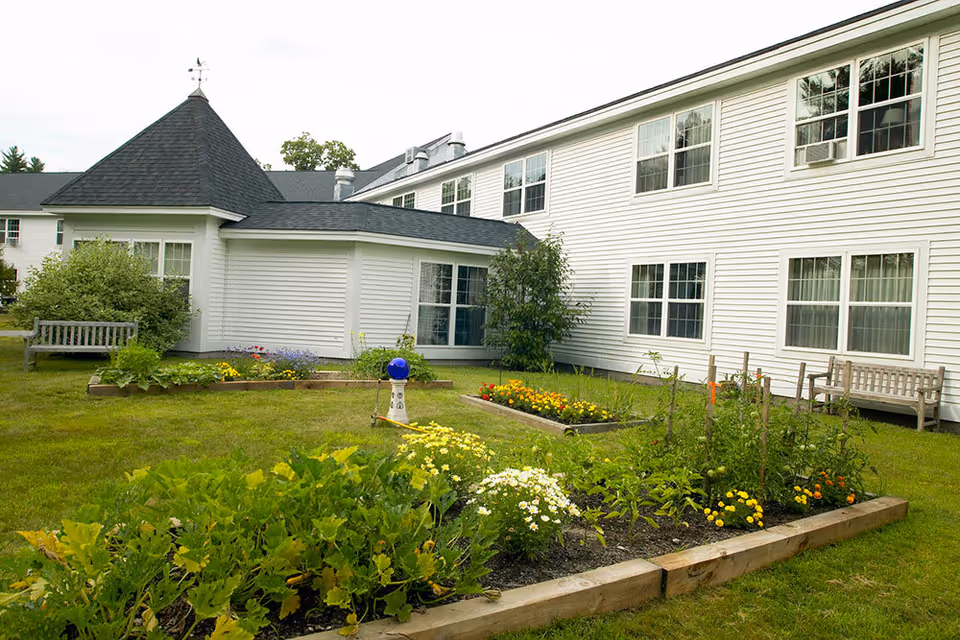 White two-story building with a grassy courtyard, raised garden beds and wooden benches.
