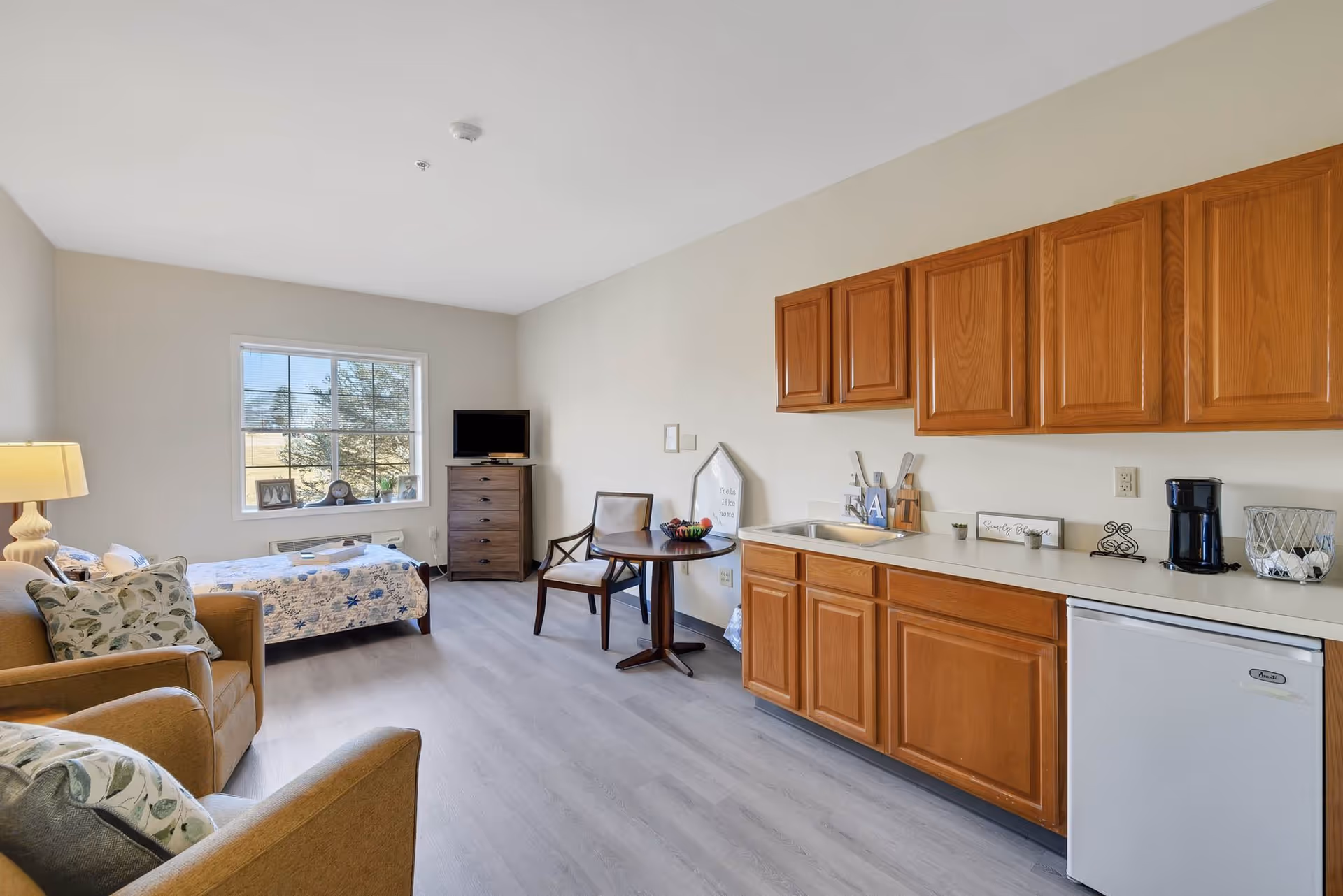 A bright senior living studio apartment featuring a bed with a floral quilt near a window, a small wooden table with two chairs, a TV on a dresser, two armchairs with patterned pillows, and a kitchenette with wooden cabinets, a sink, a coffee maker, and a mini fridge.