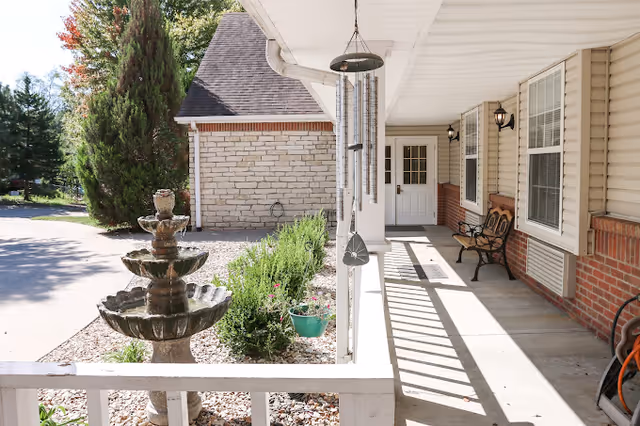 Covered front porch with a three-tier stone fountain, wind chimes, a bench, and a walkway leading to a white entry door.