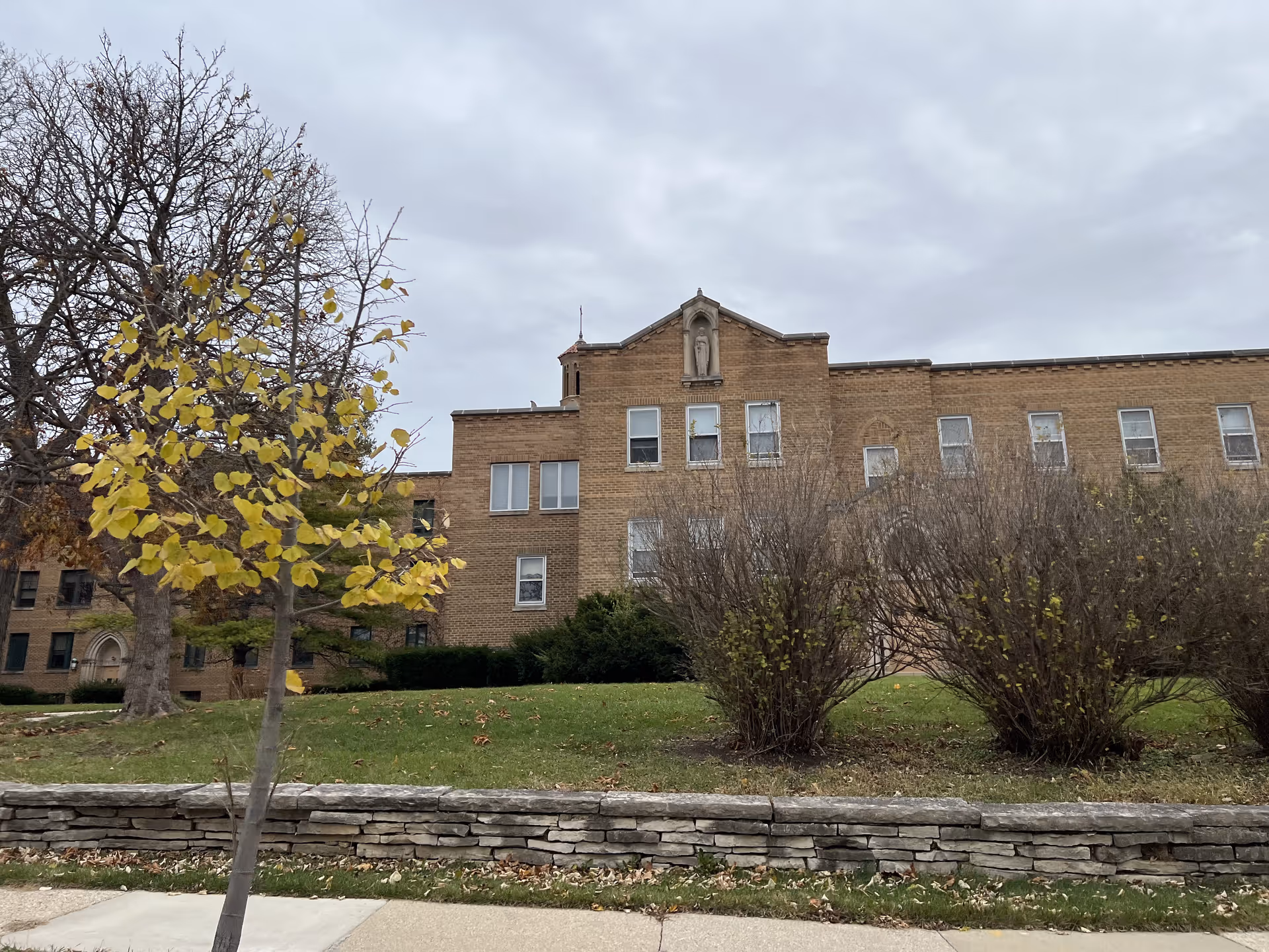 Exterior view of a large brick building with multiple windows, a small statue in a niche near the roof, and a grassy area with bushes and a young tree with yellow leaves in front. The sky is overcast.