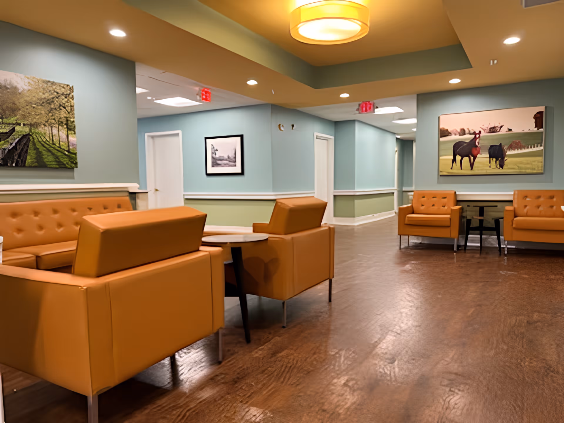 A seating area in a senior living facility with orange cushioned chairs and sofas arranged around small tables. The walls are painted in light blue and green with white trim, and decorated with framed artwork. The ceiling has recessed lighting and a large circular light fixture. The floor is wooden, and there are several white doors along the hallway.