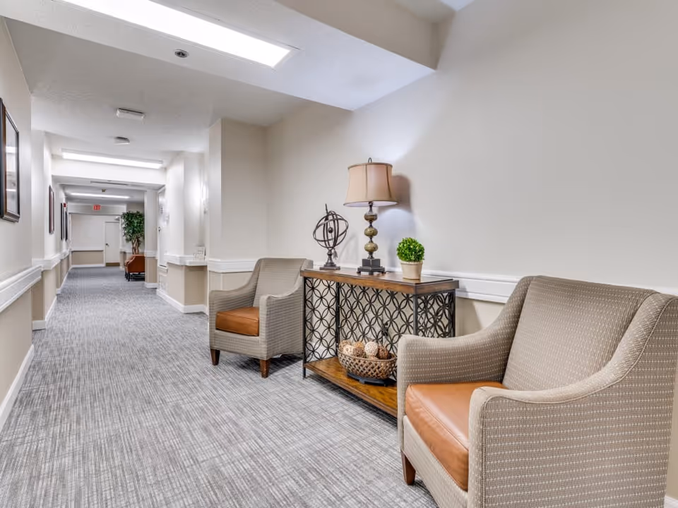 A well-lit hallway in a senior living facility with two beige armchairs featuring brown cushions placed next to a decorative console table. The table holds a lamp, a small potted plant, and a decorative orb. The hallway has light-colored walls, carpeted flooring, framed pictures on the walls, and a potted plant visible further down the corridor.