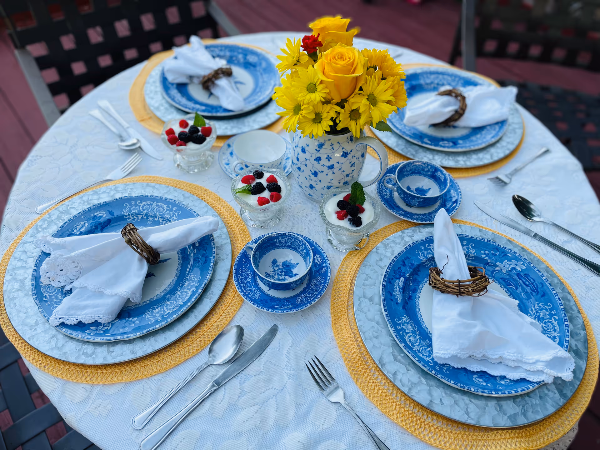 Outdoor round table set for four with blue-and-white china, white napkins in wicker rings, a yellow floral centerpiece, and small berry desserts.