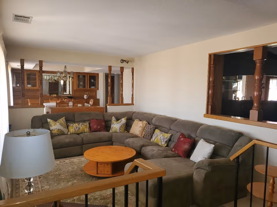 A cozy living room featuring a large gray sectional sofa adorned with various patterned throw pillows. In front of the sofa is a round wooden coffee table. To the left, there is a floor lamp with a white shade. The room has light-colored walls and a carpeted floor. Wooden railings and columns separate the living room from the dining area, which contains a wooden dining table and chairs, and a chandelier hanging above the table.