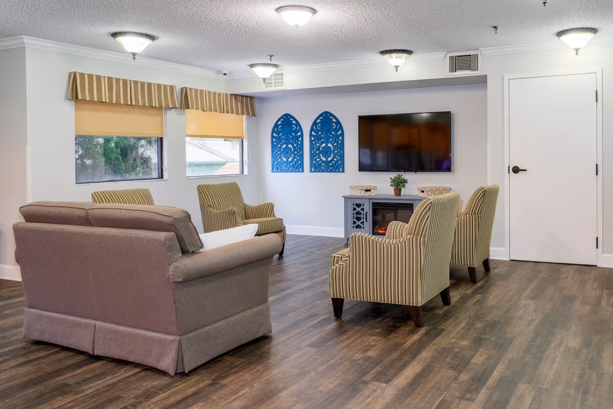 A cozy living room with a brown sofa and four striped armchairs arranged around a wall-mounted flat screen TV above a small electric fireplace. The room has wood flooring, two windows with beige roller shades and striped valances, and two decorative blue wall hangings. The ceiling has multiple light fixtures and there is a white door on the right side.