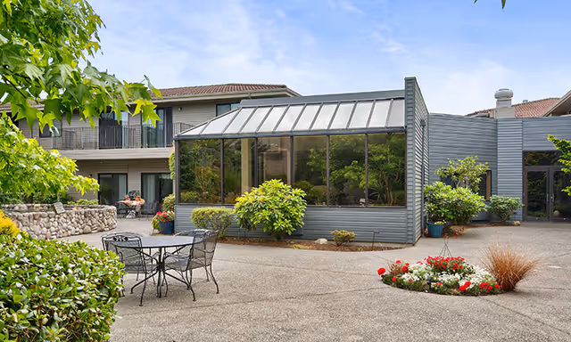 Outdoor courtyard area of a retirement and assisted living facility featuring a glass-enclosed sunroom, metal patio table with chairs, various green shrubs and plants, a small flower bed with red and white flowers, and a two-story building with balconies in the background under a partly cloudy sky.