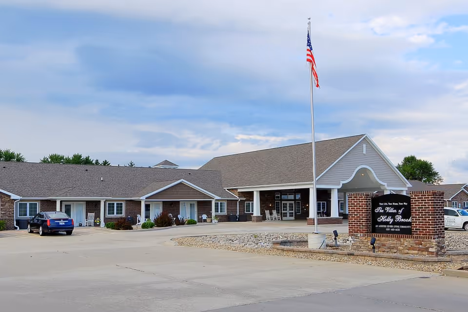 Exterior view of Villas of Holly Brook Chatham, a single-story brick building with a large covered entrance, an American flag on a flagpole, and a sign displaying the facility's name. There are a few cars parked in front and some greenery around the building.