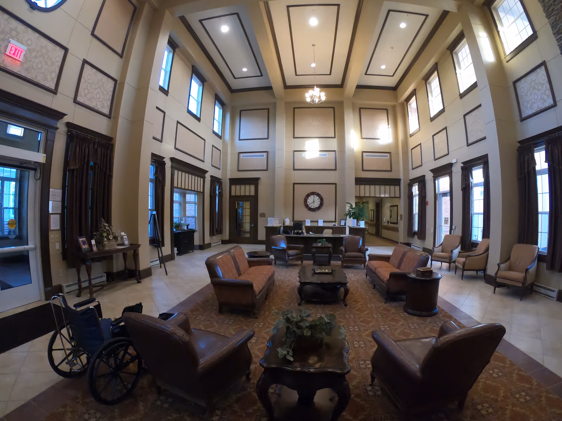 A spacious and well-lit nursing home lobby with high ceilings and multiple tall windows. The room is furnished with several leather sofas and armchairs arranged around coffee tables on a large patterned rug. There is a wheelchair near the entrance, a clock on the far wall above a reception desk, and decorative plants and framed pictures on side tables. The walls have decorative paneling and the ceiling features recessed lighting and a chandelier.