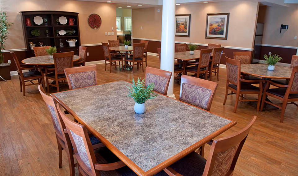 Well-lit dining area with multiple tables and chairs, potted centerpieces, wood floors, and framed artwork on the walls.