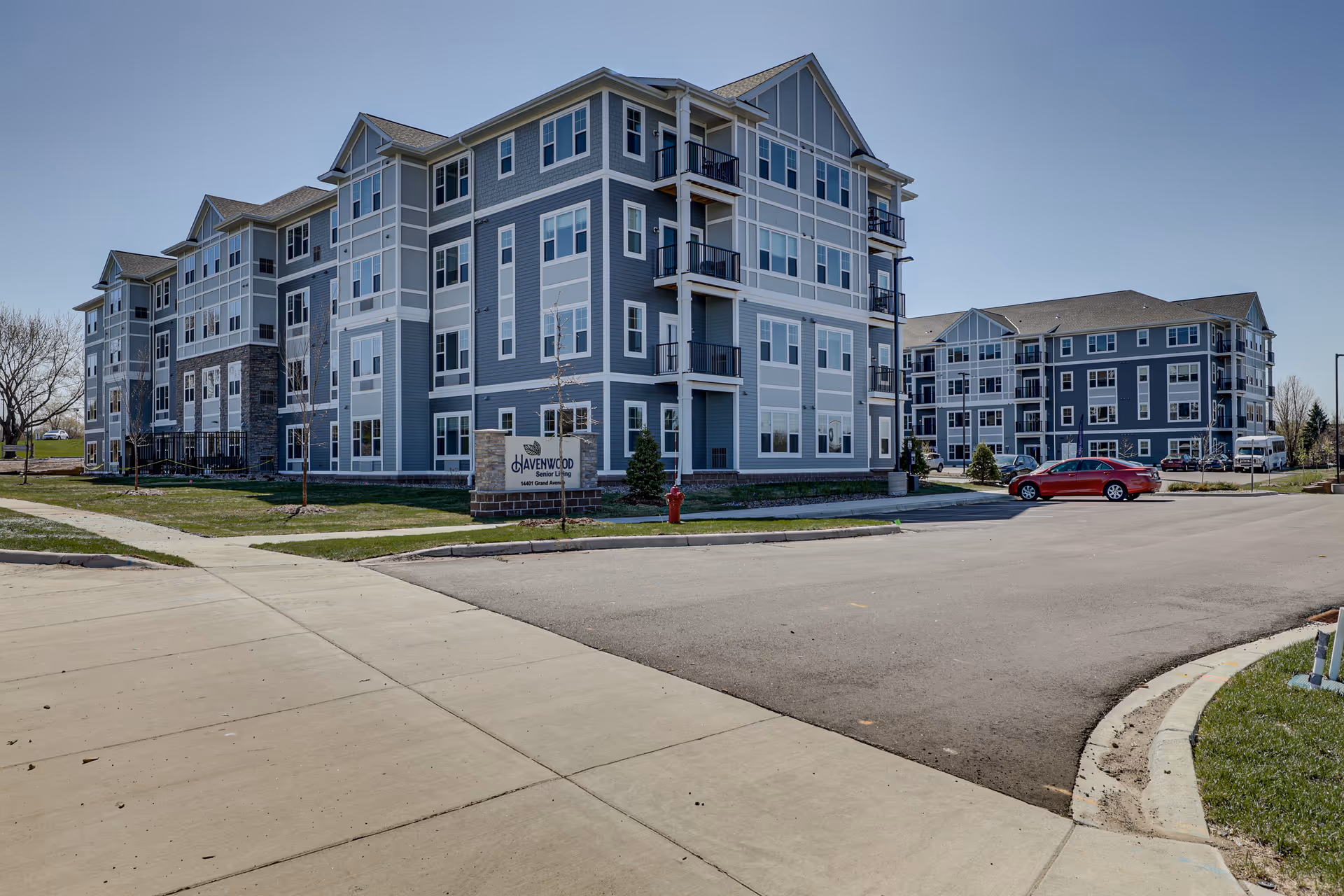 Exterior view of Havenwood of Burnsville, a multi-story senior living facility with blue and gray siding, multiple windows, balconies, and a parking area with cars. The building is surrounded by a sidewalk, grass, and a clear blue sky.