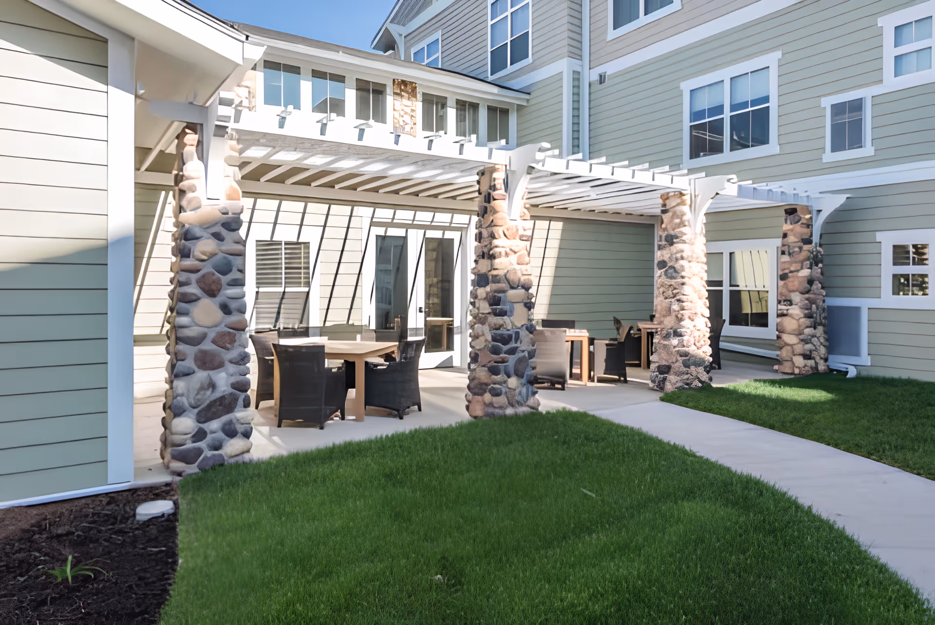 Outdoor patio area at Heartis Peoria with stone pillars supporting white pergolas, several tables and chairs arranged for seating, surrounded by green grass and adjacent to the building with multiple windows.