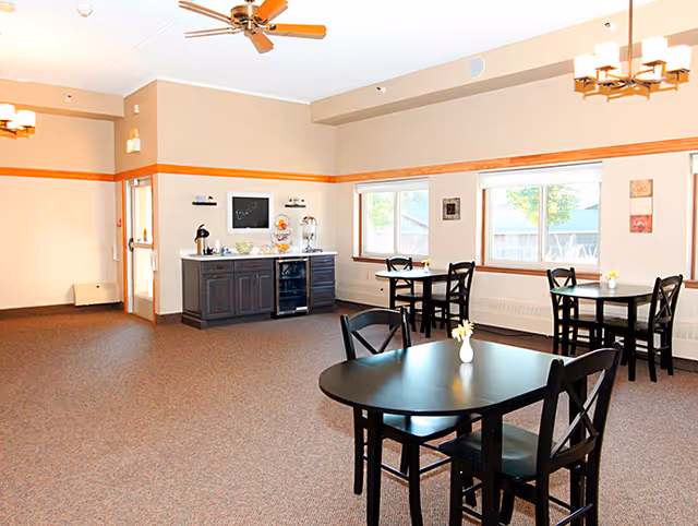A bright dining room with several black tables and chairs, each table decorated with a small vase holding a yellow flower. The room has beige walls with wooden trim, large windows letting in natural light, ceiling fans, and a sideboard with a coffee maker and beverage dispenser.