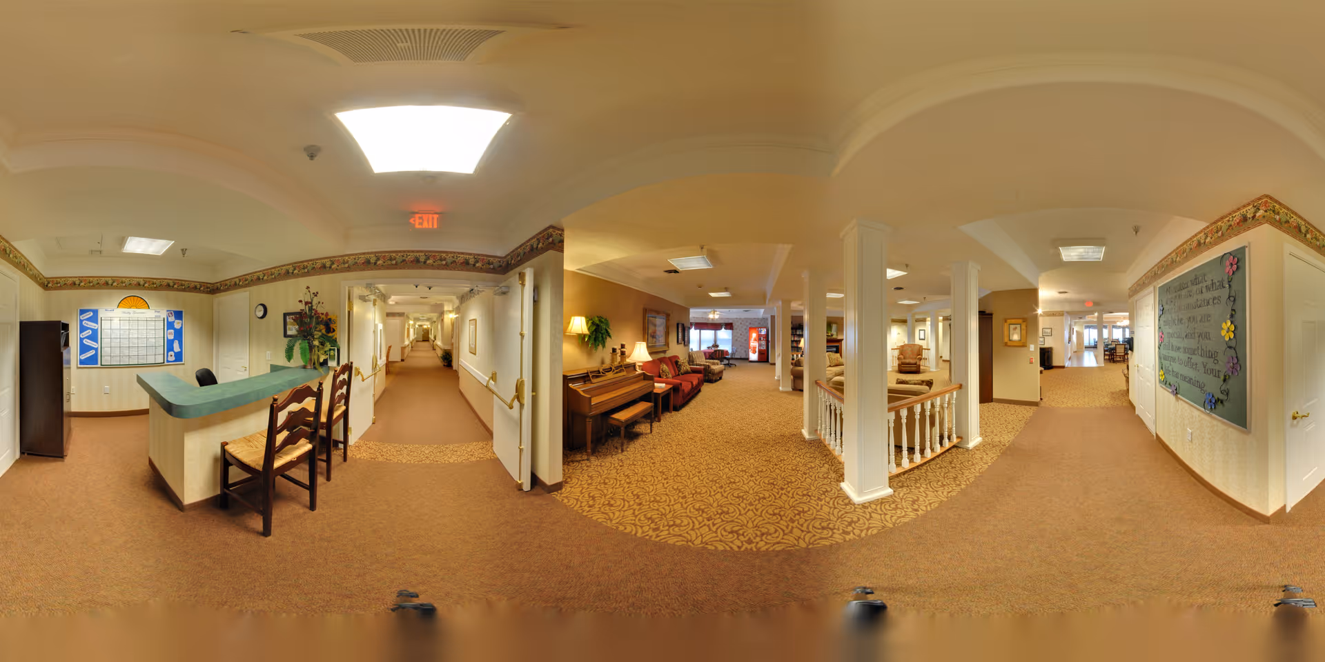 Panoramic view of an interior senior living facility hallway and common area. The scene includes a reception desk with chairs, a long corridor with handrails, a piano with seating, decorative plants, and a bulletin board with information. The space is warmly lit with carpeted floors and light-colored walls.