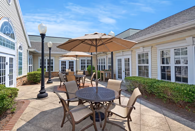 Sunlit courtyard patio with round metal tables, umbrellas, chairs, lamp posts, and a single-story building with many windows and doors.