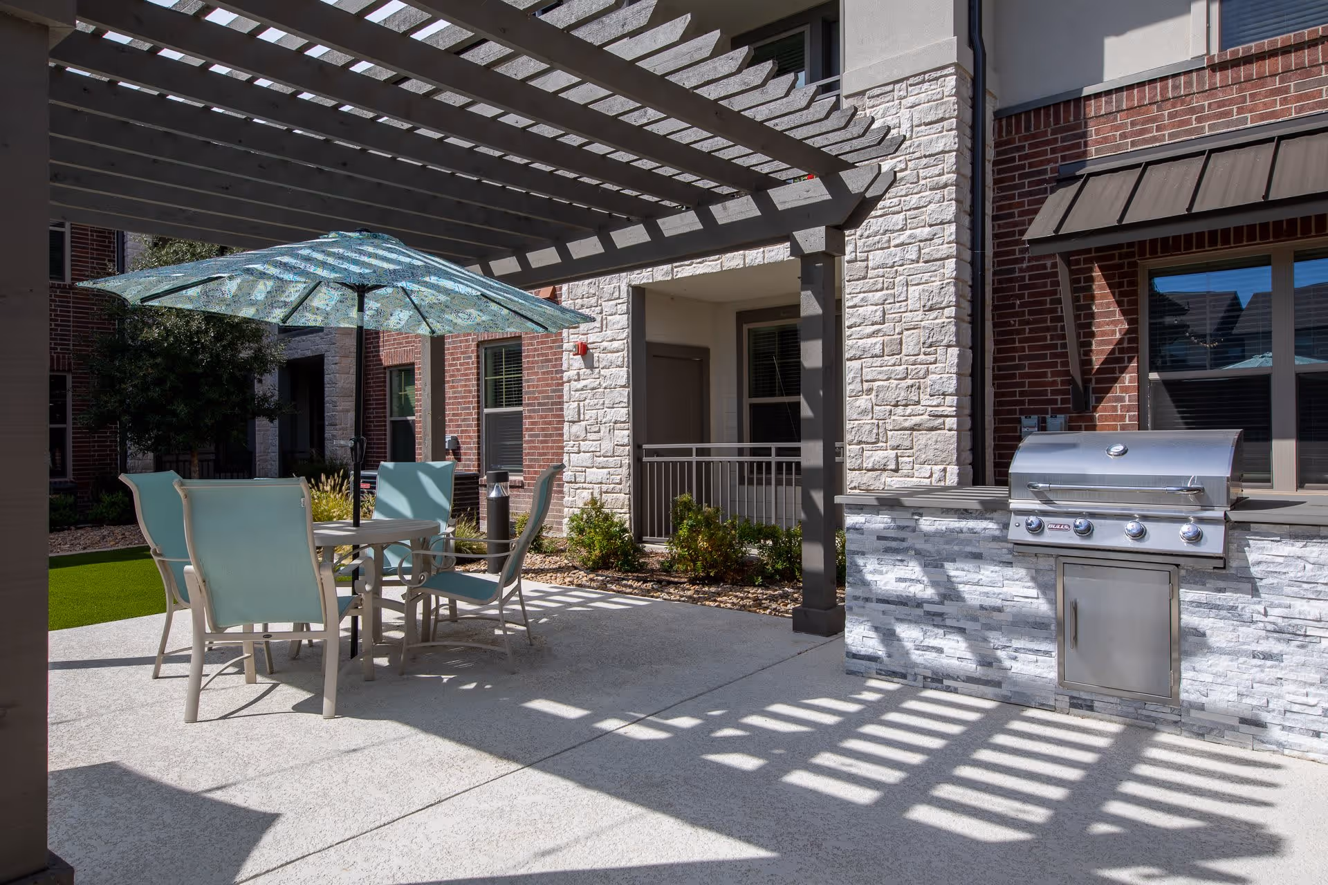 Outdoor patio area with a pergola casting shadows on the ground, a round table with four light blue chairs and a blue patterned umbrella, and a stainless steel built-in grill against a stone and brick building exterior.