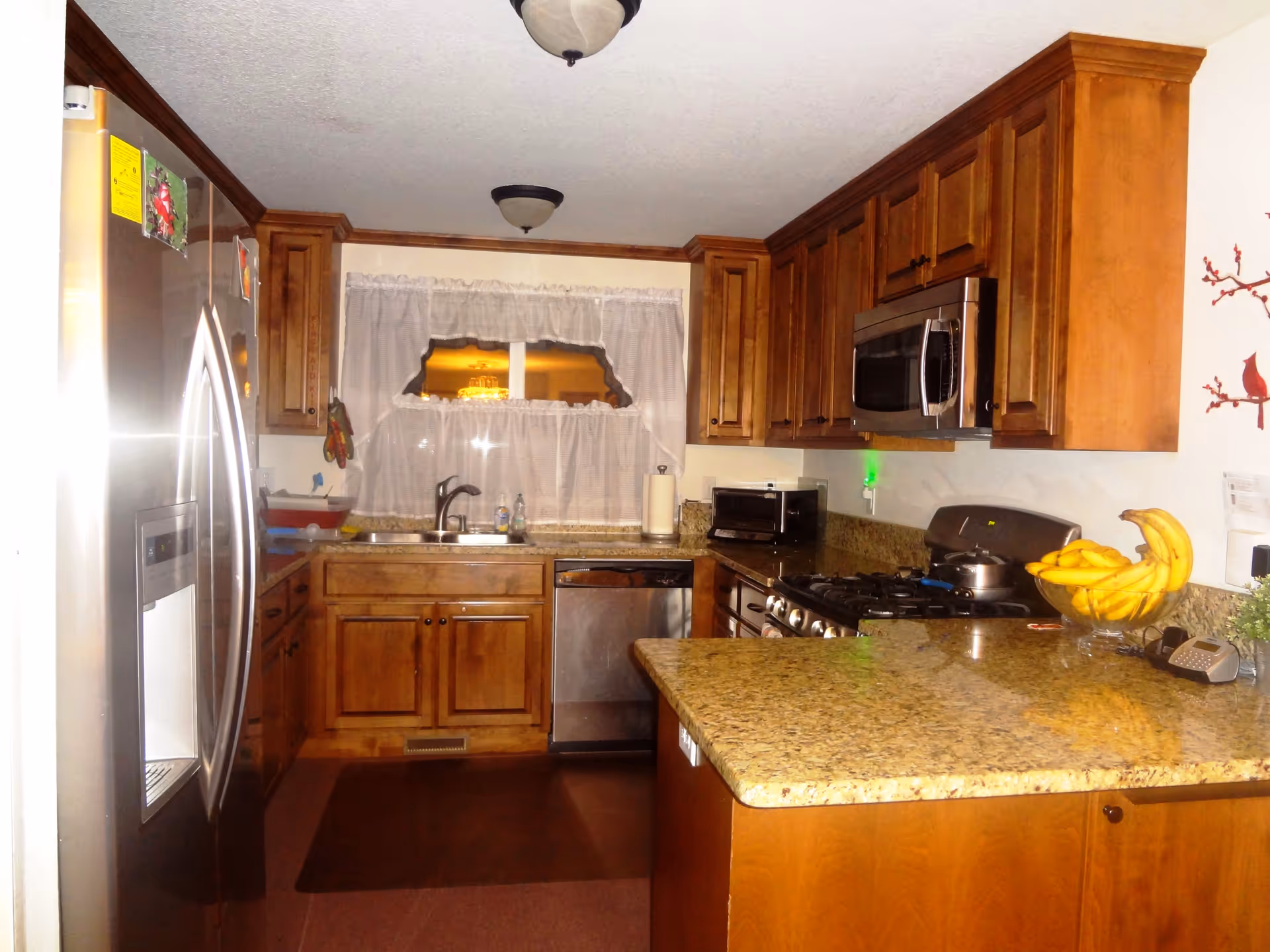 A kitchen with wooden cabinets, granite countertops, a stainless steel refrigerator, dishwasher, microwave, and gas stove. There is a window with white curtains above the sink, a bowl of bananas on the counter, and a small toaster oven on the countertop.