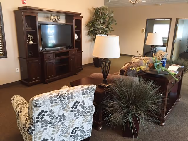 A cozy living room area featuring a large dark wood entertainment center with a flat-screen TV, a patterned armchair, a brown sofa, two table lamps, a coffee table with decorative plants, and a large potted plant in the corner. The room has beige walls and carpeted flooring.