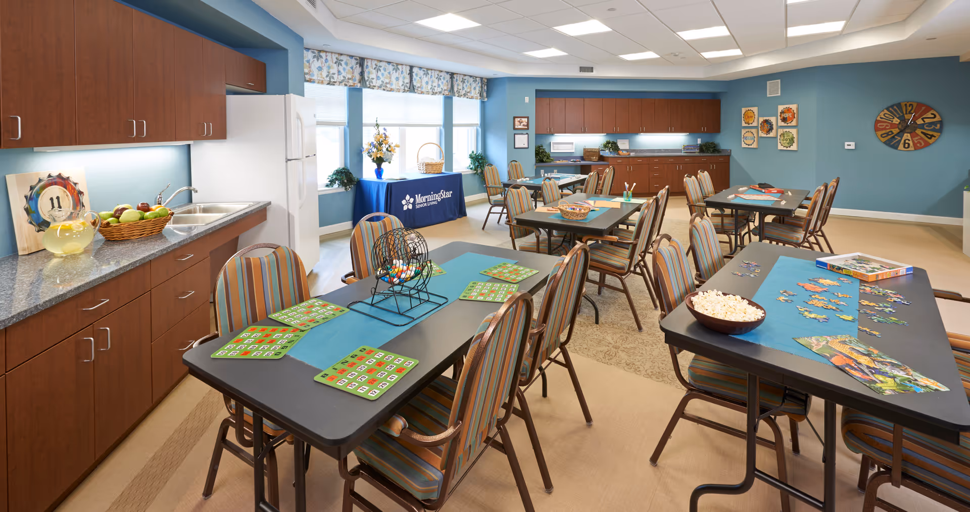 A bright and spacious activity room with multiple tables and chairs. One table has bingo cards and a bingo cage, while another table has a partially completed jigsaw puzzle and a bowl of popcorn. The room features blue walls, wooden cabinets, a refrigerator, and large windows with floral valances. A table with a MorningStar Assisted Living & Memory Care at Mountain Shadows branded cloth is positioned near the windows, decorated with a vase of flowers and a basket.