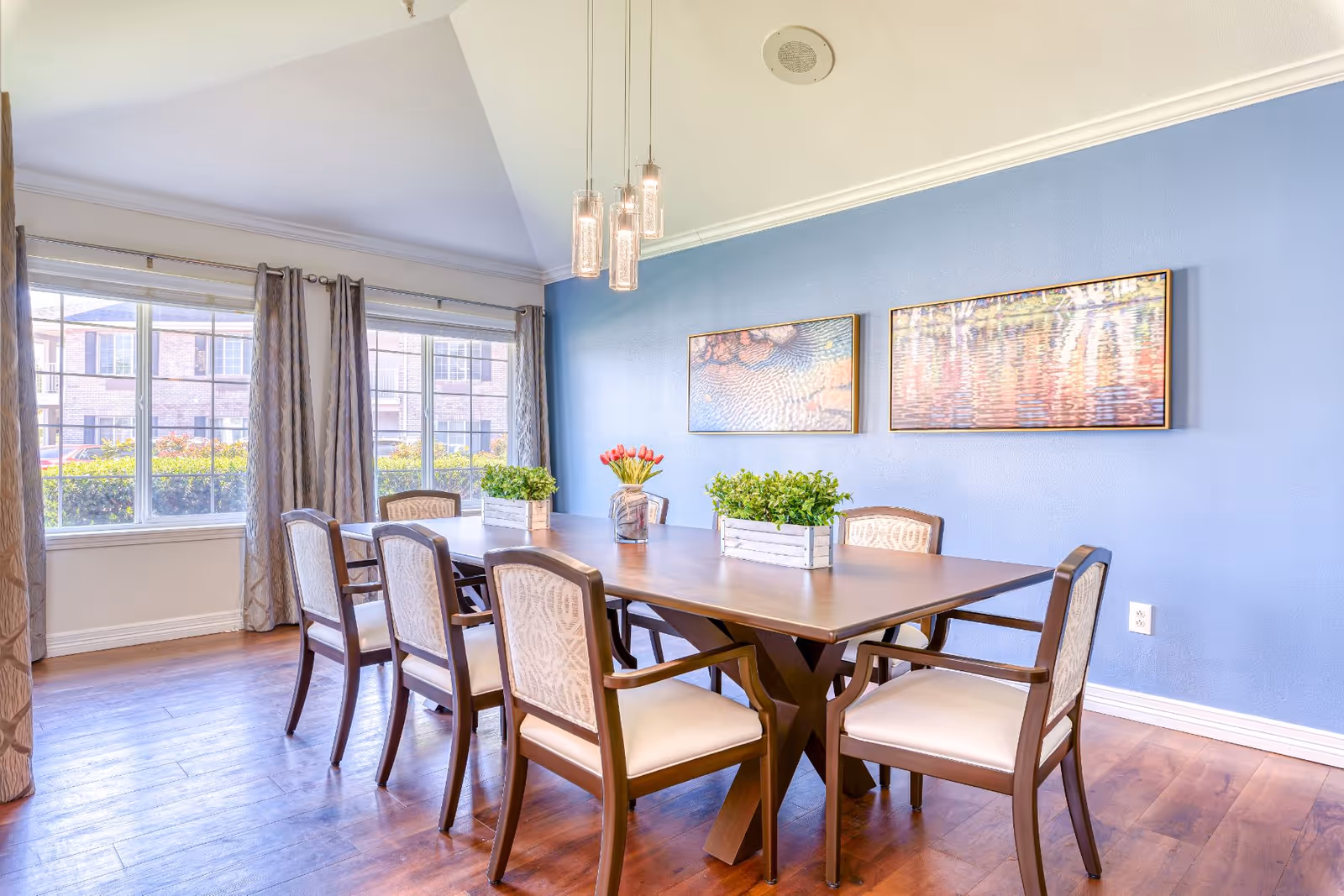A bright dining room with a wooden table surrounded by eight cushioned chairs. The walls are painted blue and decorated with two framed pictures. Three pendant lights hang from the ceiling above the table. Large windows with curtains allow natural light to fill the room, and there are two small green plants and a vase with red tulips on the table.