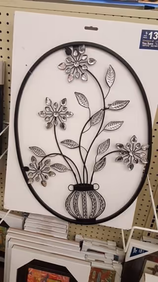 Oval metal wall art showing a vase with flowering stems and mirrored petals displayed on pegboard shelving.