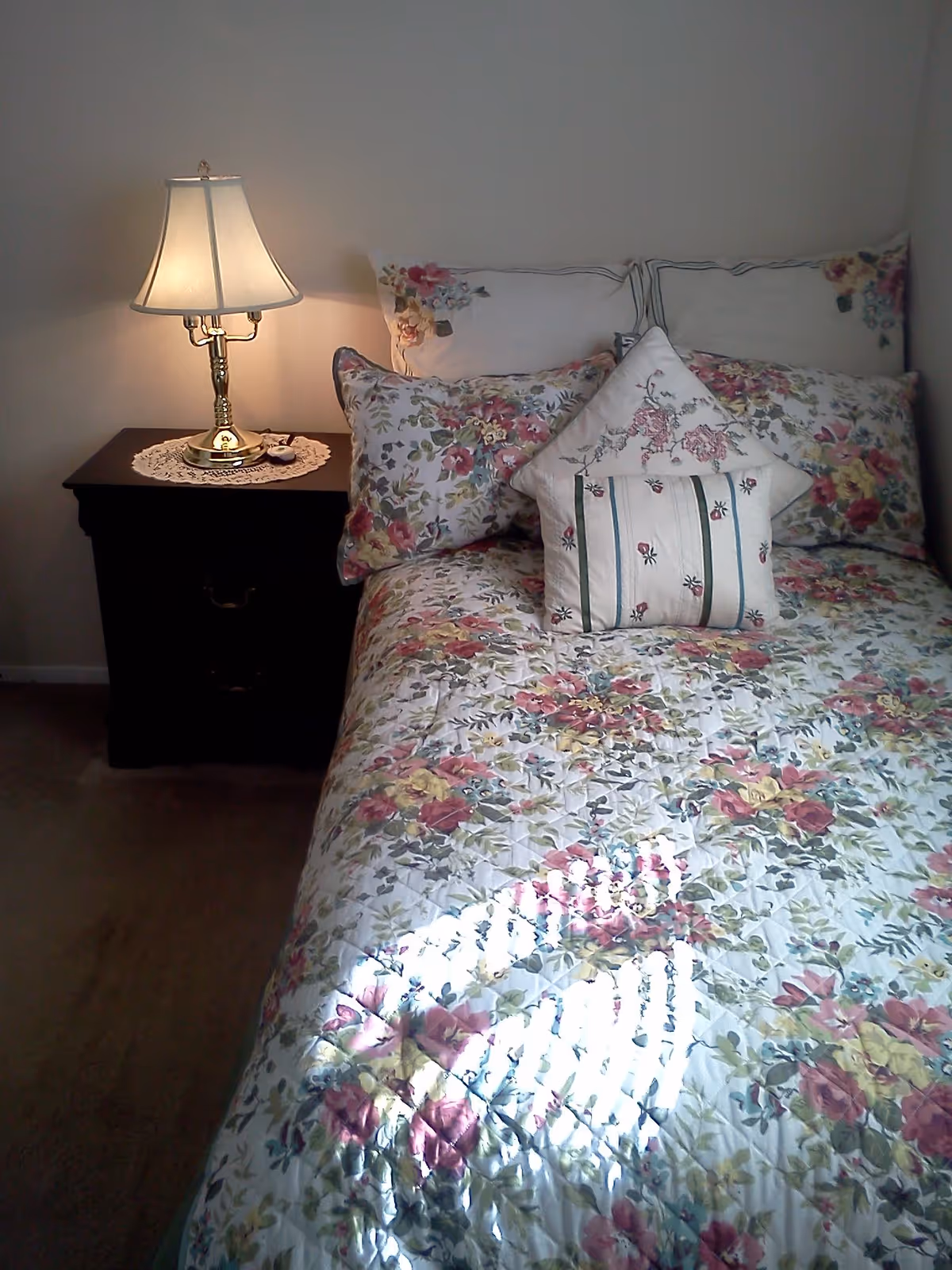A neatly made bed with floral bedding and decorative pillows beside a nightstand with a lit table lamp.