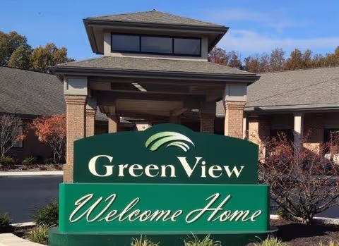 Entrance of GreenView Senior Assisted Living facility with a large green sign that reads 'GreenView Welcome Home' in front of a brick building under a clear blue sky.