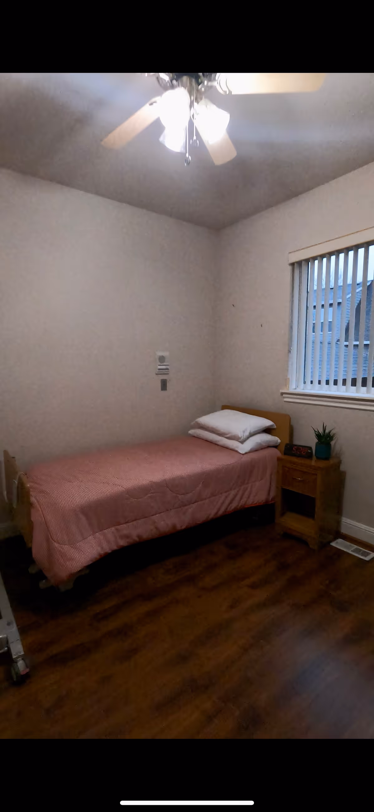 A small bedroom with a single bed covered in a pink bedspread and two white pillows. Next to the bed is a wooden nightstand with a small plant and a digital alarm clock. The room has wooden flooring, a ceiling fan with lights, and a window with vertical blinds.