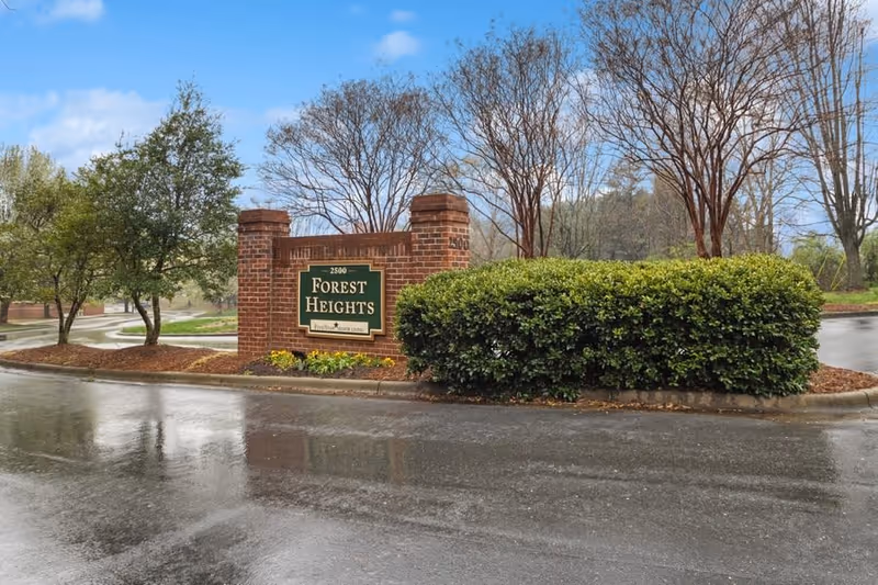 Brick entrance sign for Forest Heights with surrounding bushes and trees on a wet road under a cloudy sky.