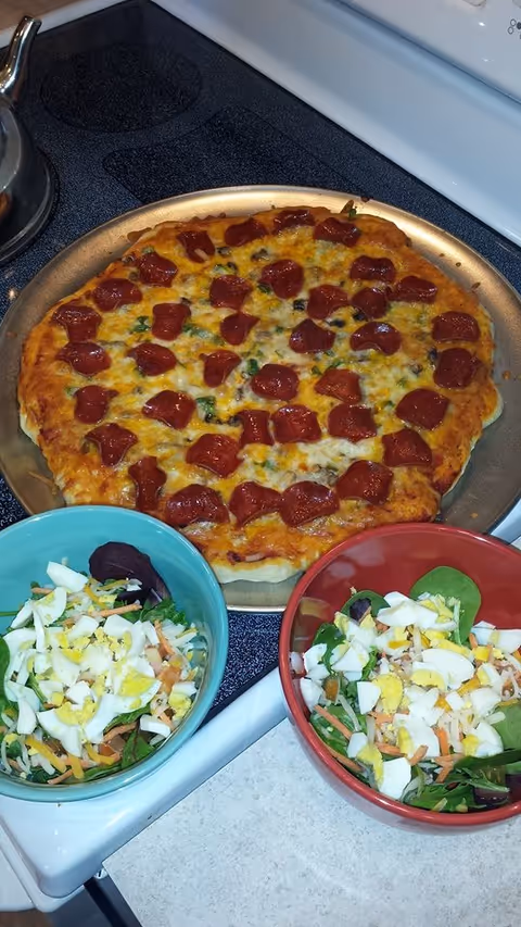 A freshly baked pepperoni pizza on a round metal tray placed on a stovetop. In front of the pizza, there are two bowls of salad, one in a blue bowl and the other in a red bowl, both containing chopped boiled eggs, shredded cheese, and leafy greens.
