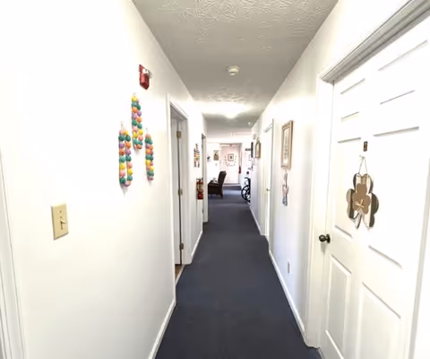 Well-lit carpeted hallway in an assisted living facility with doors and wall decorations leading toward a sitting area.