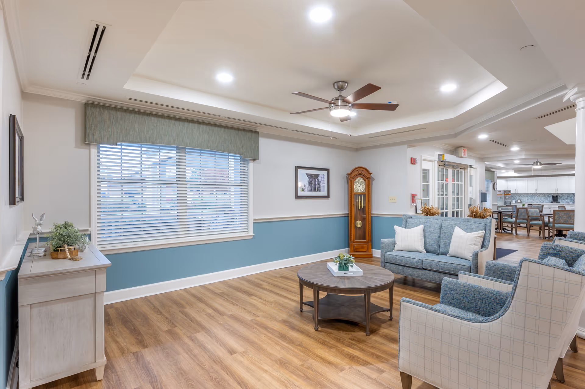 A bright and spacious living room area in Brookdale Finneytown featuring wood flooring, a ceiling fan with lights, a large window with blinds and a valance, a wooden grandfather clock, a round coffee table with a small plant centerpiece, a blue upholstered loveseat with white pillows, and two matching armchairs. In the background, there is a dining area with tables and chairs and a kitchen with white cabinetry.