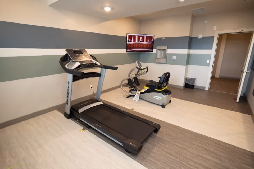 A small exercise room with a treadmill and a recumbent exercise bike. The walls have horizontal stripes in shades of gray and green. A flat-screen TV is mounted on the wall above the exercise bike. There is an open door leading to another room and a trash bin near the wall.