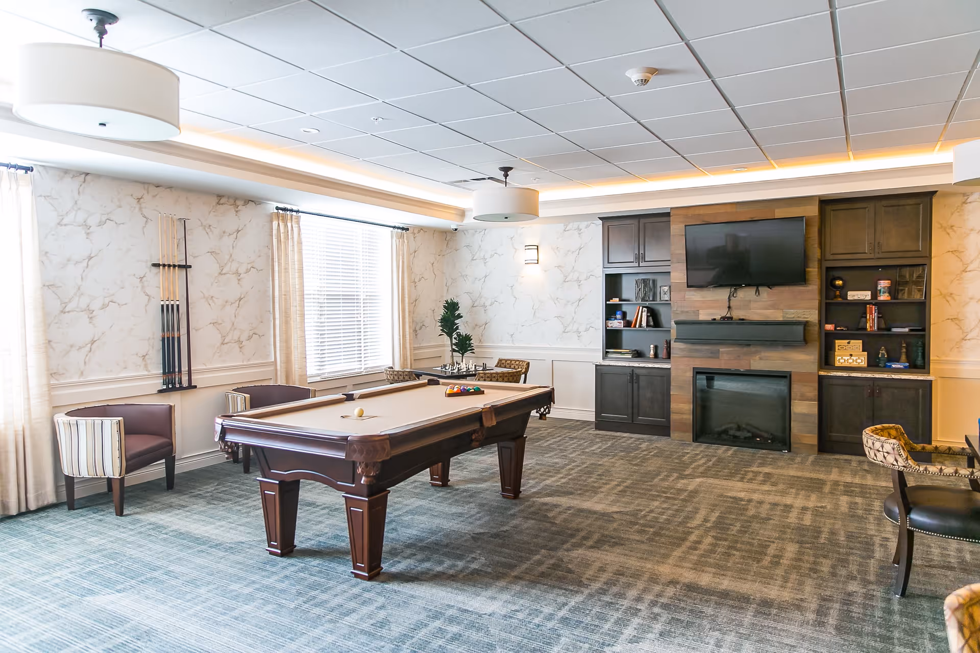 A well-lit recreational room with a pool table in the center, surrounded by chairs and a rack holding pool cues on the wall. The room features a carpeted floor, a large window with curtains, built-in dark wood cabinets, a wall-mounted TV above a modern fireplace, and decorative wallpaper.