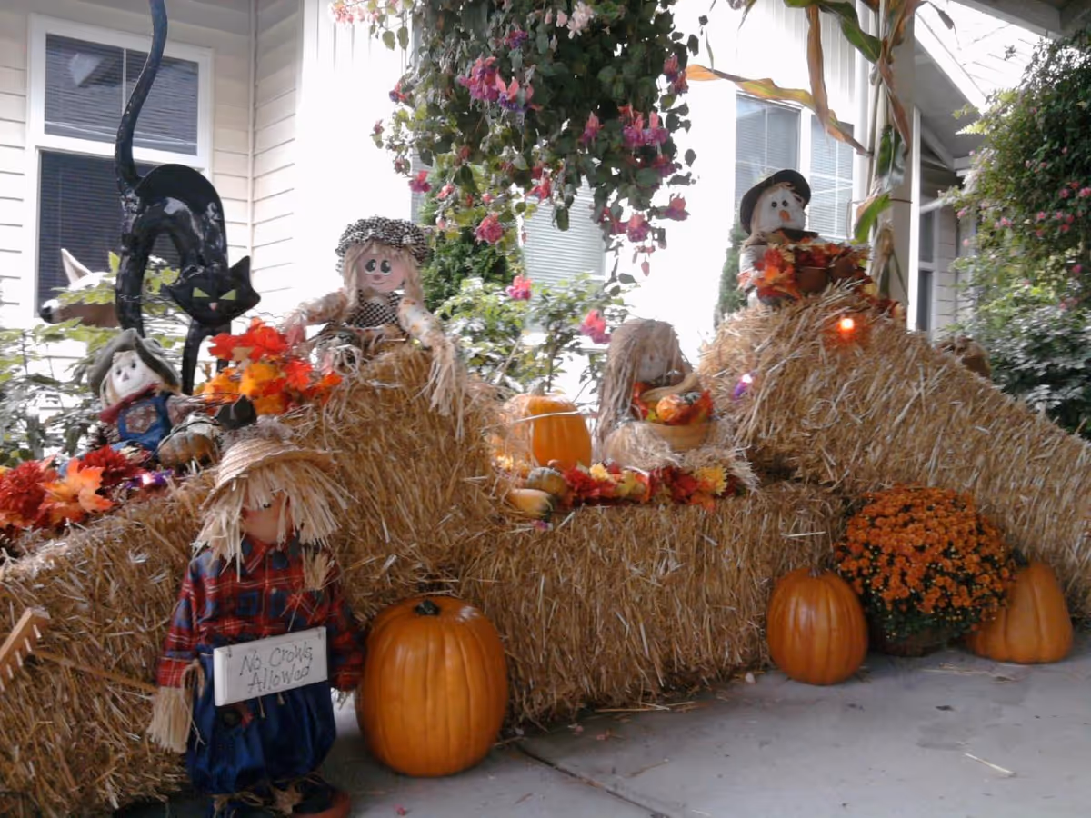 A festive autumn display featuring several scarecrow dolls, pumpkins, hay bales, and fall flowers arranged on a porch with hanging flower baskets and a house window in the background.