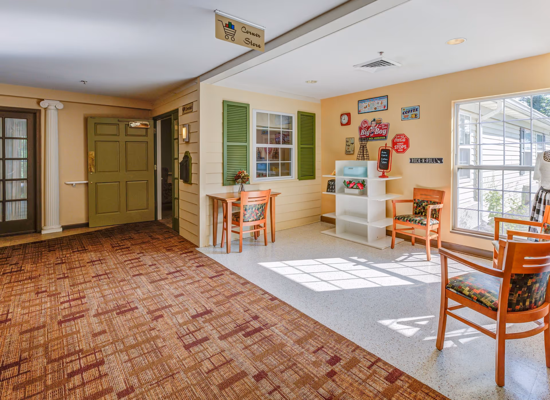 A bright interior corner of a senior living facility featuring a small seating area with wooden chairs upholstered in patterned fabric, a small table with a flower vase, and a white shelving unit. The walls are decorated with vintage-style signs and a large window lets in natural light. The floor transitions from patterned carpet to a light-colored tile. A sign above reads 'Corner Store'.