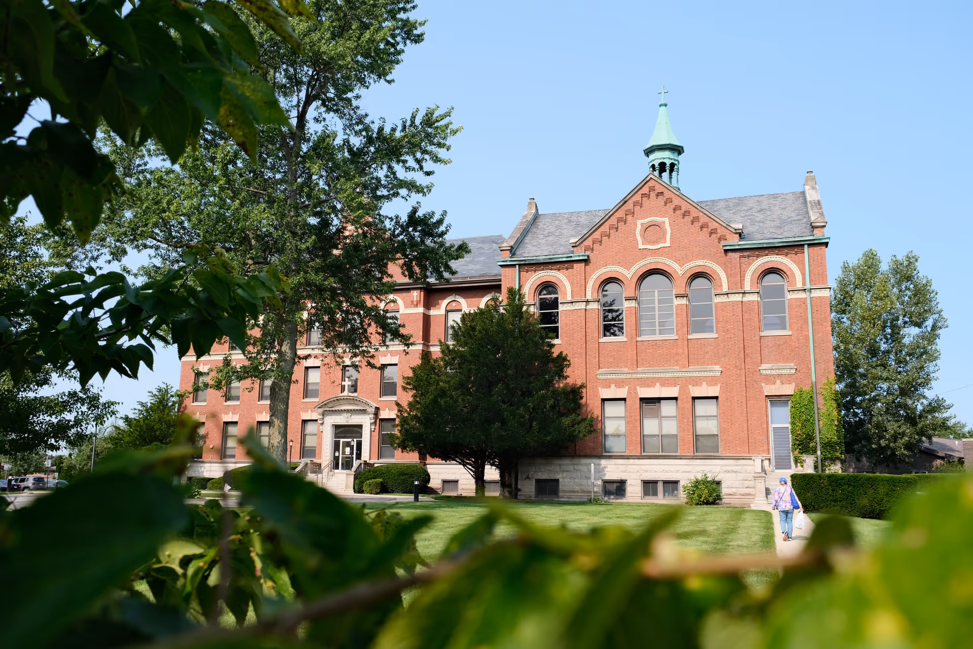 A large, historic red brick building with arched windows and a green steeple, surrounded by green trees and a well-maintained lawn. A person is walking on the sidewalk towards the building entrance on a clear, sunny day.