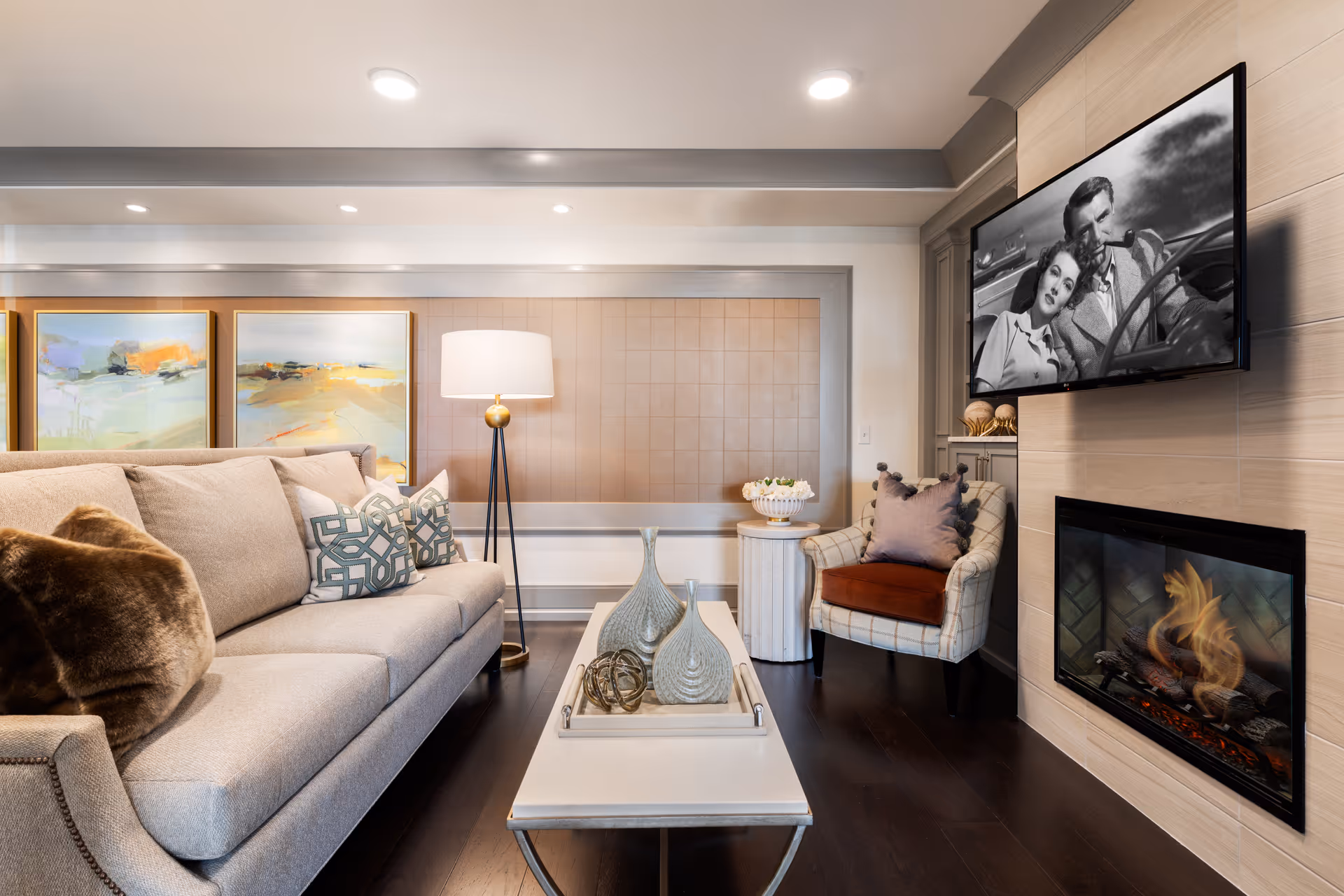 A cozy living room with a beige sofa adorned with decorative pillows, a modern floor lamp, a white coffee table with decorative vases, a plaid armchair with a purple pillow, a wall-mounted TV displaying a black and white movie scene, and a fireplace with a warm fire.