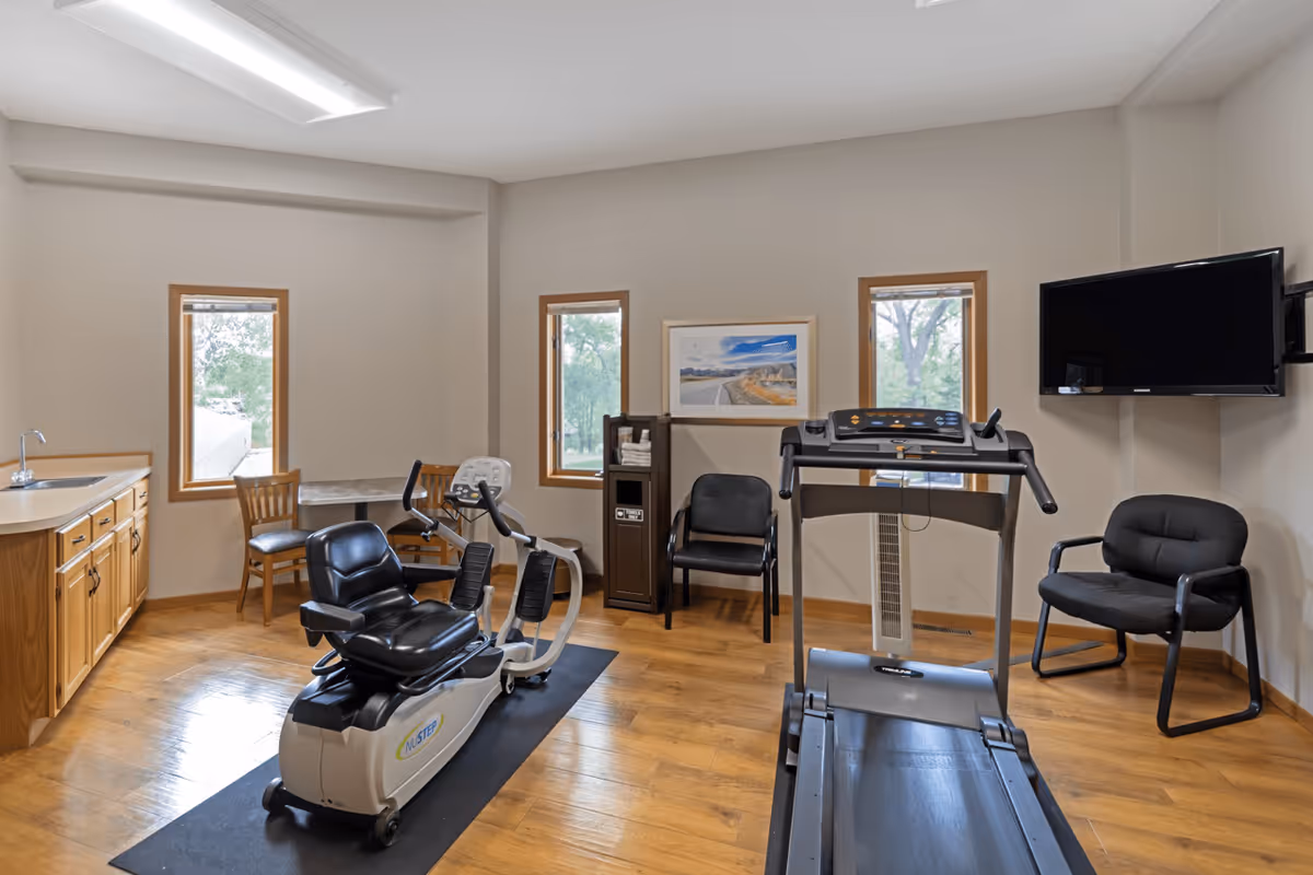 A small exercise room with wooden flooring and beige walls featuring a recumbent exercise bike and a treadmill on black mats. The room has three windows with wooden frames letting in natural light. There are two black chairs, a wooden chair, a small table, a cabinet with towels, a wall-mounted flat screen TV, and a framed landscape picture on the wall.