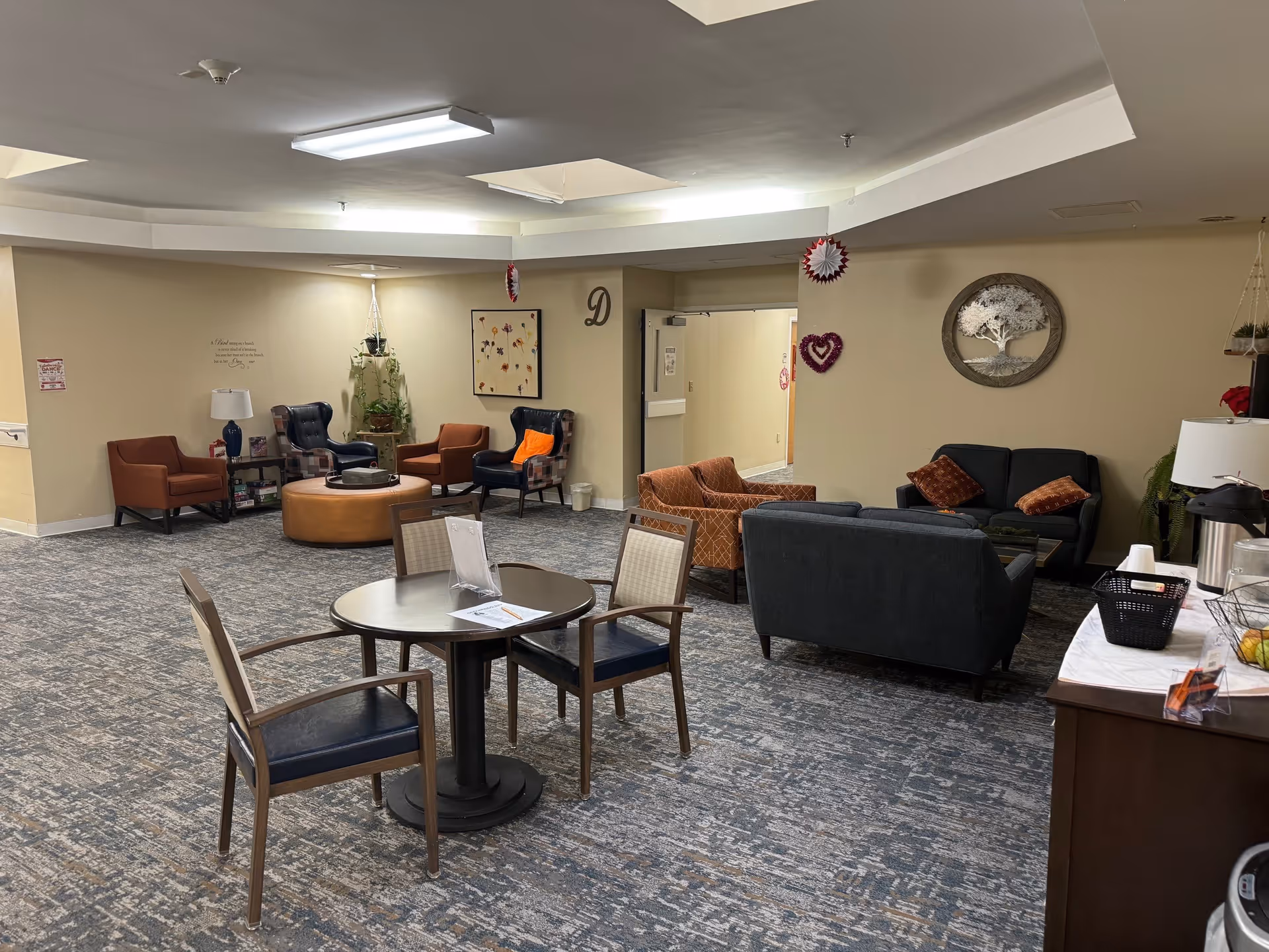 A senior living facility common area with multiple seating arrangements including armchairs, sofas, and a round table with chairs. The room has beige walls decorated with framed artwork and hanging plants. There is a sideboard with a coffee dispenser and cups on the right side. The carpet is patterned in shades of blue and gray.