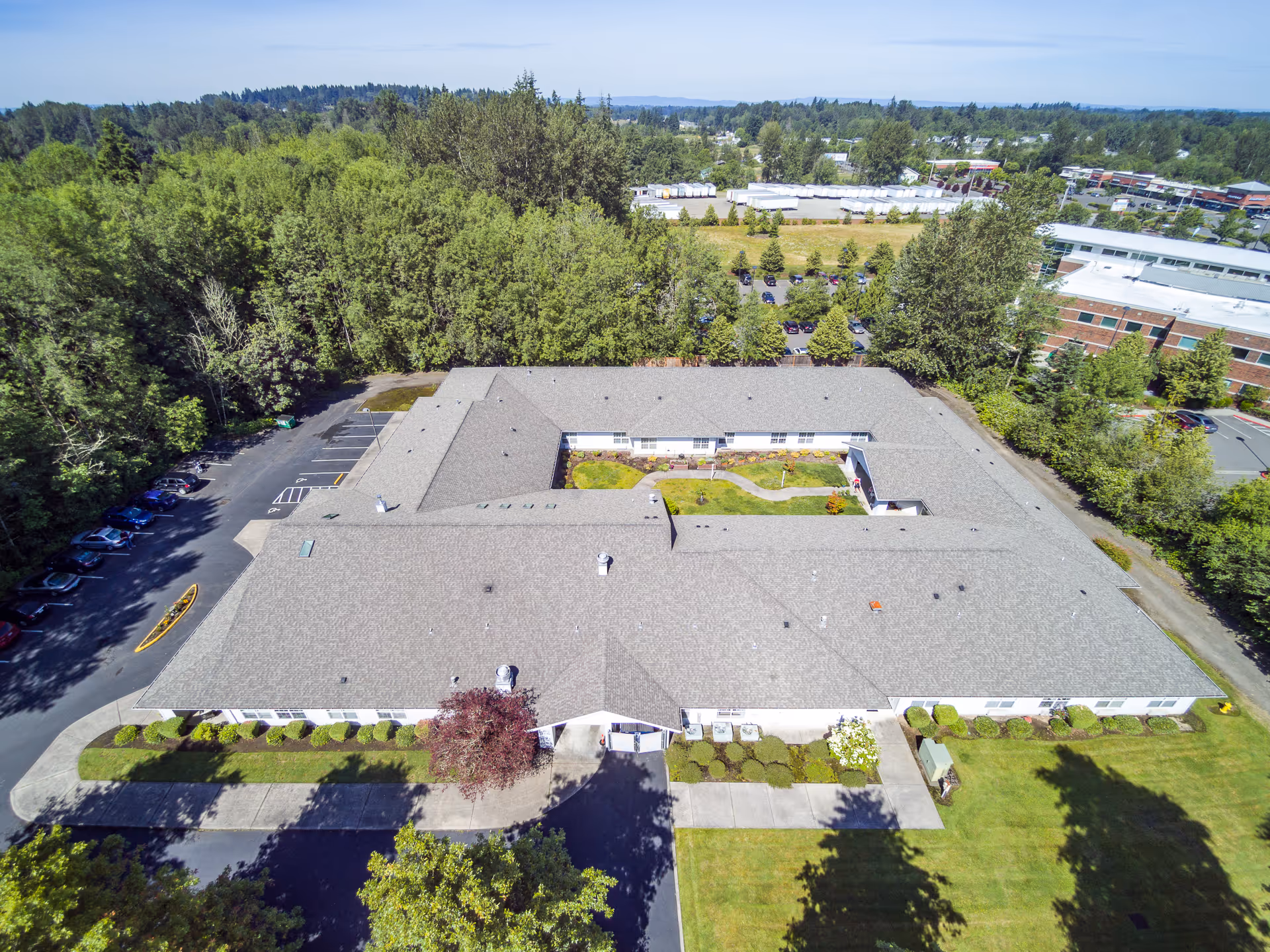 Aerial view of Creekside Place Memory Care facility showing a large, single-story building with a central courtyard. The building is surrounded by trees, parking lots, and landscaped green areas under a clear blue sky.