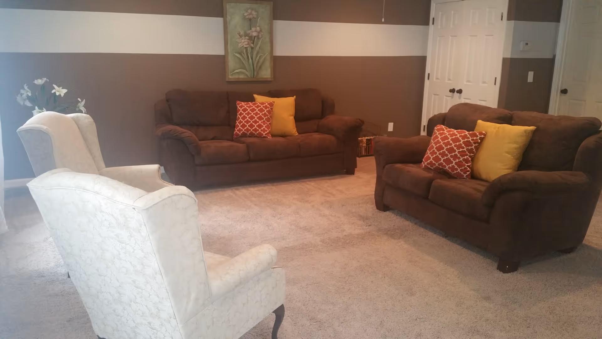 Cozy living room with brown sofas, patterned throw pillows, and two white armchairs on a carpeted floor.