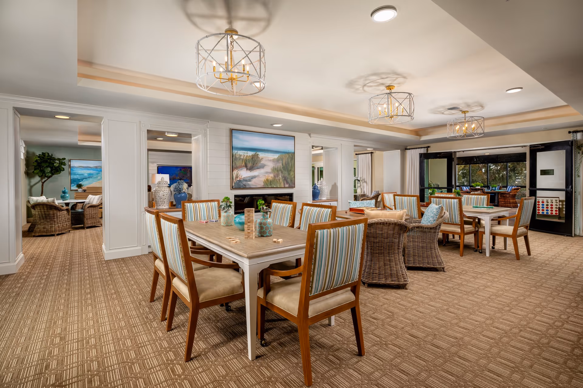 Bright communal dining and lounge area with multiple tables and chairs, wicker seating, artwork, and large windows.