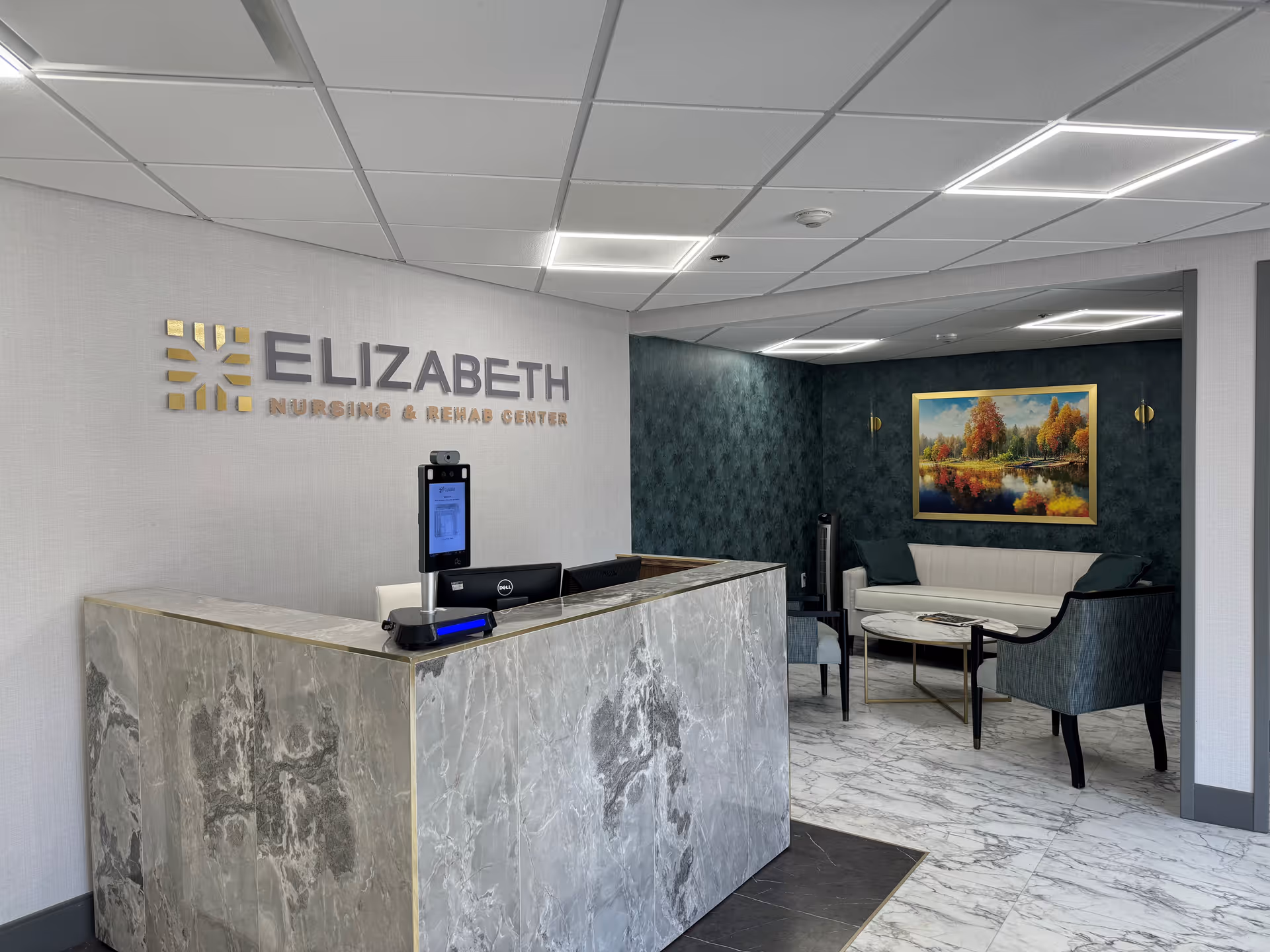 Reception area of Elizabeth Nursing and Rehabilitation Center featuring a marble reception desk with a computer and a digital check-in device. Behind the desk is a wall with the facility's name and logo. To the right, there is a seating area with a white couch, two armchairs, a round coffee table, and a framed painting of an autumn landscape on a dark green wall.