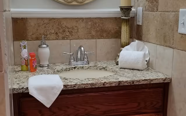 A bathroom sink with a granite countertop, a silver faucet, a soap dispenser, a few small bottles, and neatly folded white towels. The wall behind the sink is tiled with beige and brown tiles, and there is a decorative column-like candle holder on the right side.