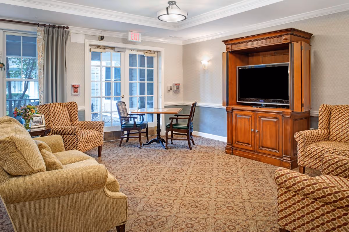 Cozy common lounge with upholstered armchairs, a wooden TV cabinet, and a small table by glass double doors.