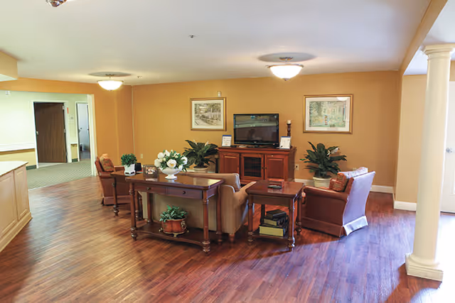 A warm communal living area with sofas and chairs arranged around a TV cabinet, wood floors, plants, and framed artwork.
