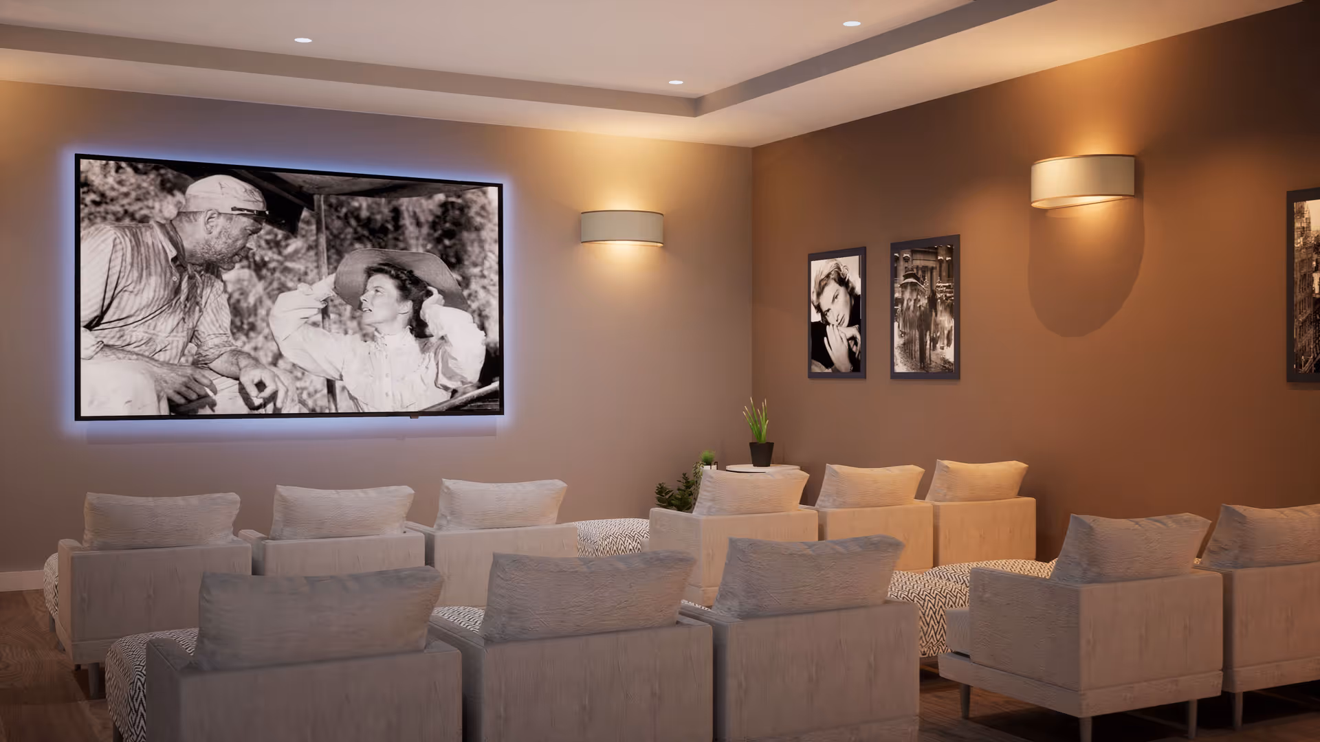 A cozy media room with multiple beige armchairs arranged in rows facing a large wall-mounted TV screen displaying a black and white movie scene. The room has warm lighting with wall sconces and framed black and white photos on the walls.