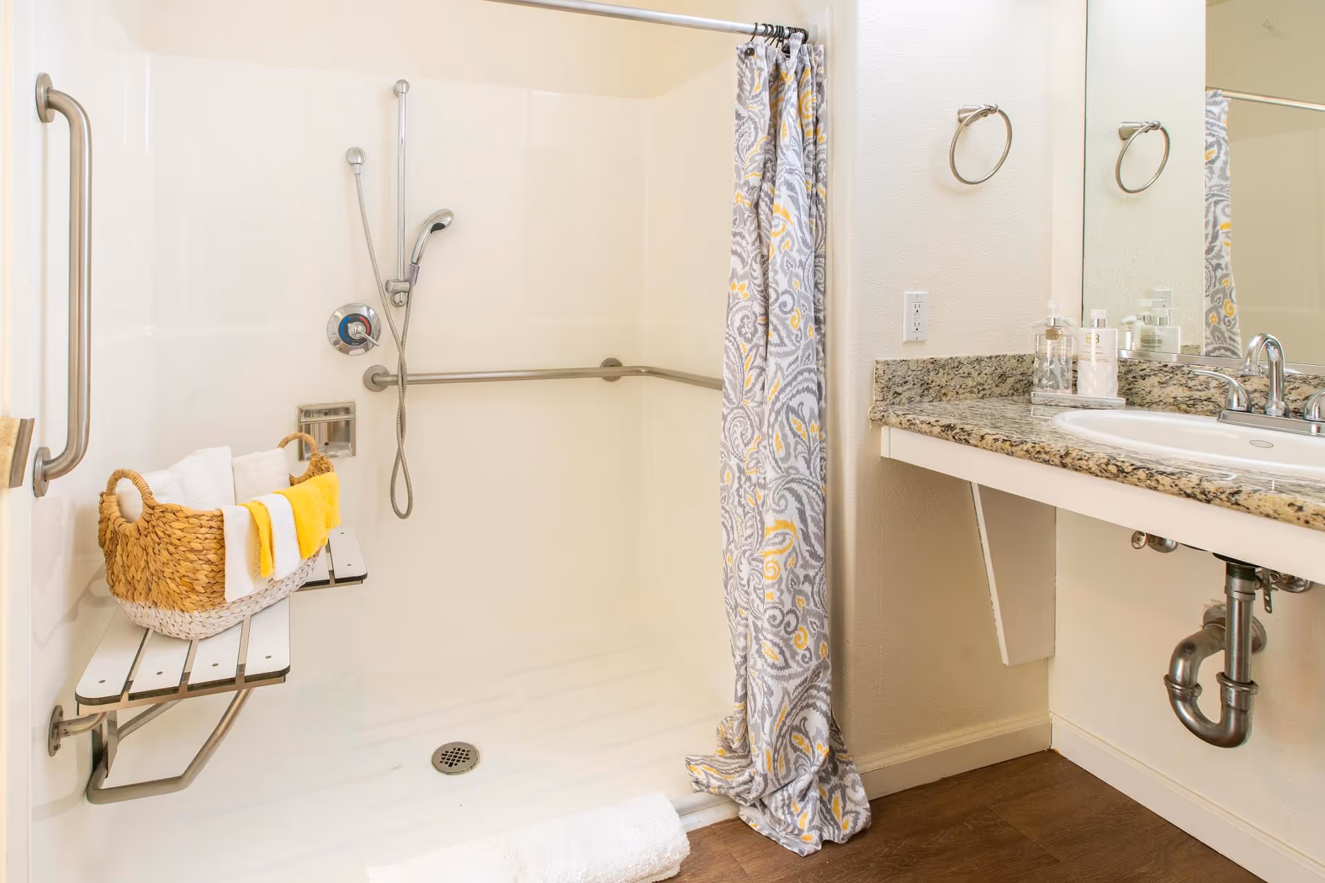 A clean and accessible bathroom shower area with a fold-down bench holding a basket of towels, grab bars on the walls, a handheld showerhead, and a patterned shower curtain. To the right is a granite countertop with a sink, soap dispensers, and a large mirror above.