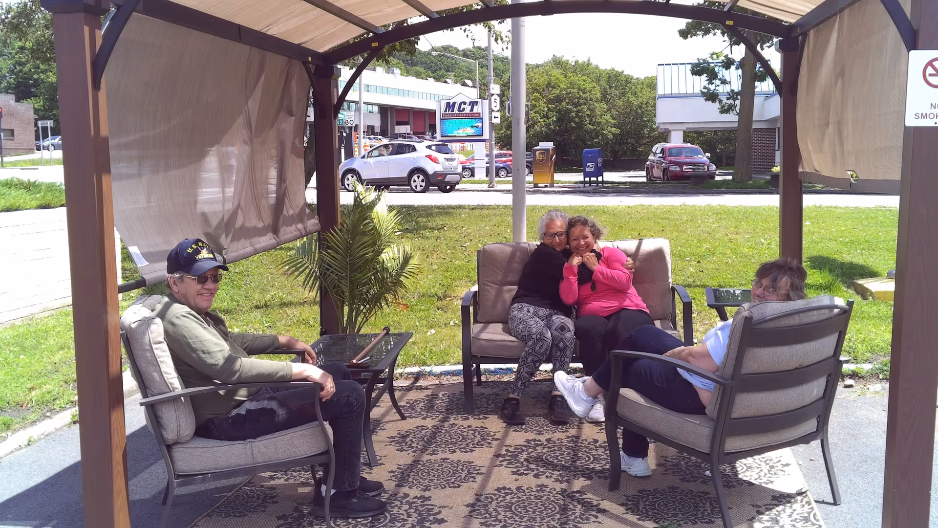 Four people sitting and chatting under a shaded outdoor gazebo with cushioned chairs and a rug.